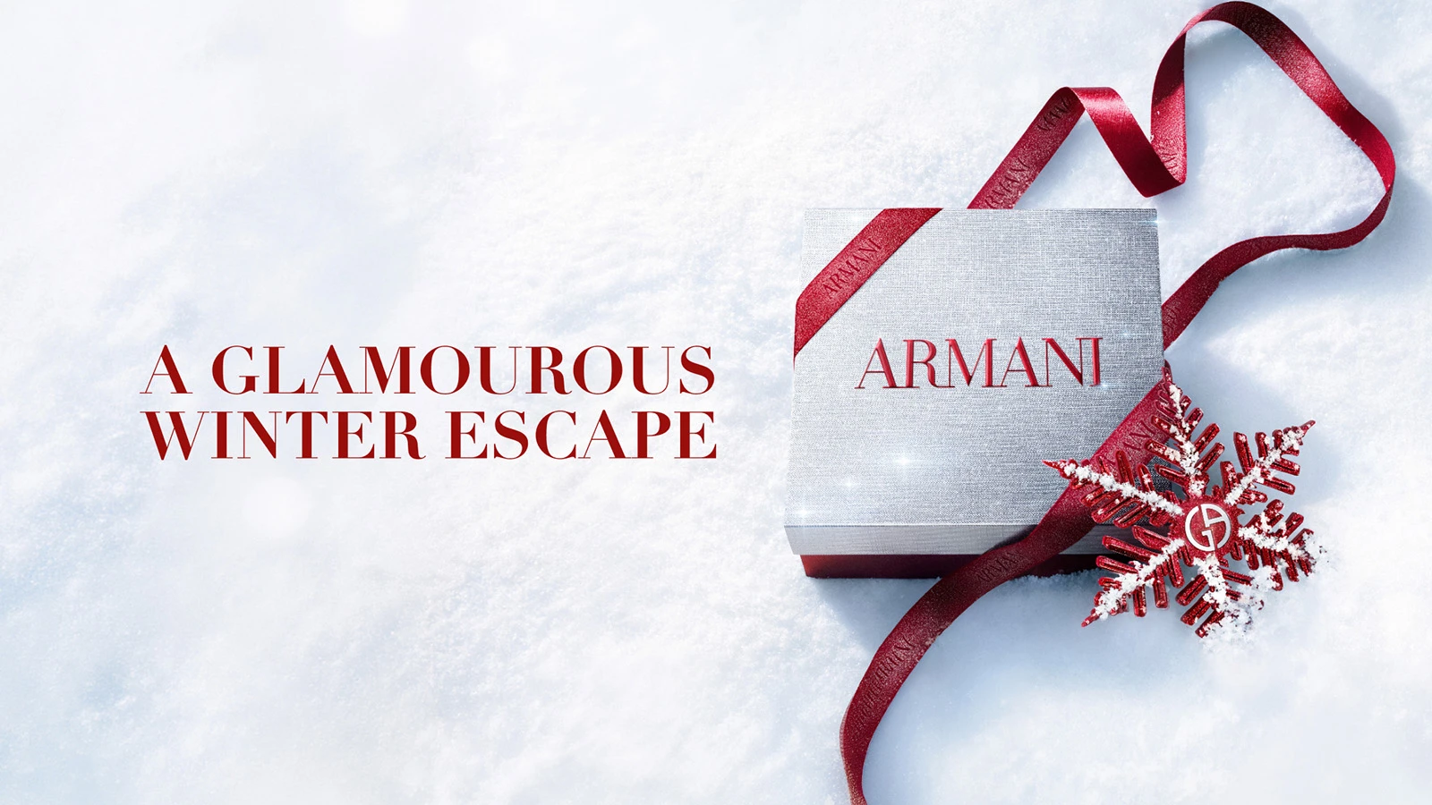 Tekst: 'EN GLAMOURØS VINTERFLUGT'. Armani gaveæske med logo, rødt bånd og snefnug på sne.