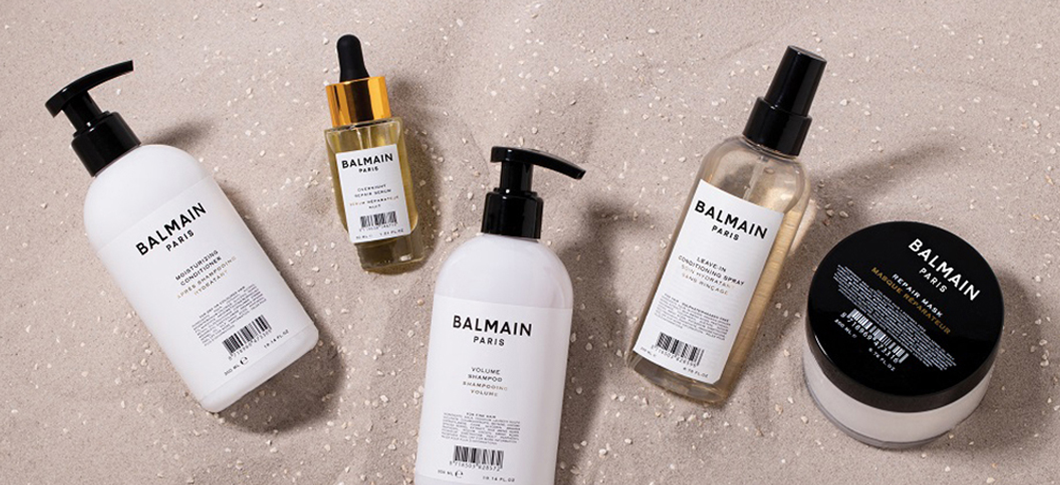 Her ses et billede med flere hårplejeprodukter fra Balmain Paris placeret på en lys, sandfarvet overflade. På produkterne står teksten “BALMAIN PARIS”, og de inkluderer blandt andet shampoo, conditioner, olie og hårmaske i hvide, gennemsigtige og sorte emballager.