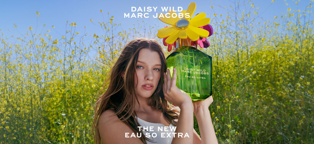 Her ses et billede med en person placeret i et blomsterfyldt landskab, hvor vedkommende holder en stor grøn parfumeflakon med blomsterformet låg. Øverst står teksten “DAISY WILD MARC JACOBS”, og nederst står “THE NEW EAU SO EXTRA”.