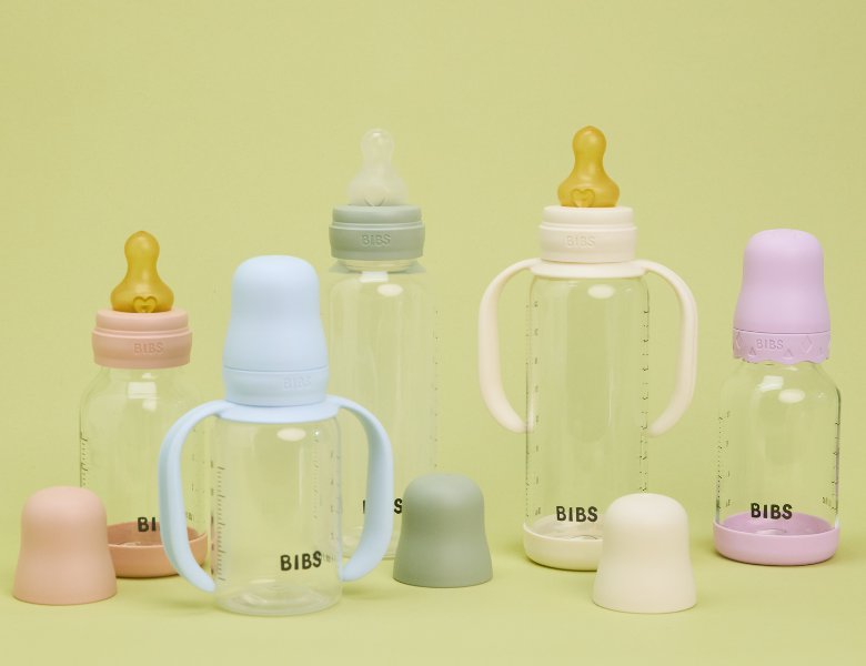 Fem BIBS babyflasker og tudkopper i pastelfarver står samlet på lysegrøn baggrund med tilhørende låg foran.