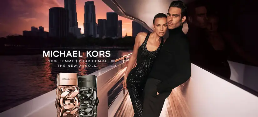 Her ses en kvinde og en mand beklædt i sort på en båd. Til venstre for dem ses to Michael Kors produkter, hvor baggrunden er en storby i solnedgang. 