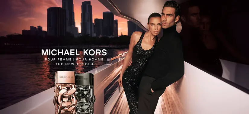 Her ses en kvinde og en mand beklædt i sort på en båd. Til venstre for dem ses to Michael Kors produkter, hvor baggrunden er en storby i solnedgang. 