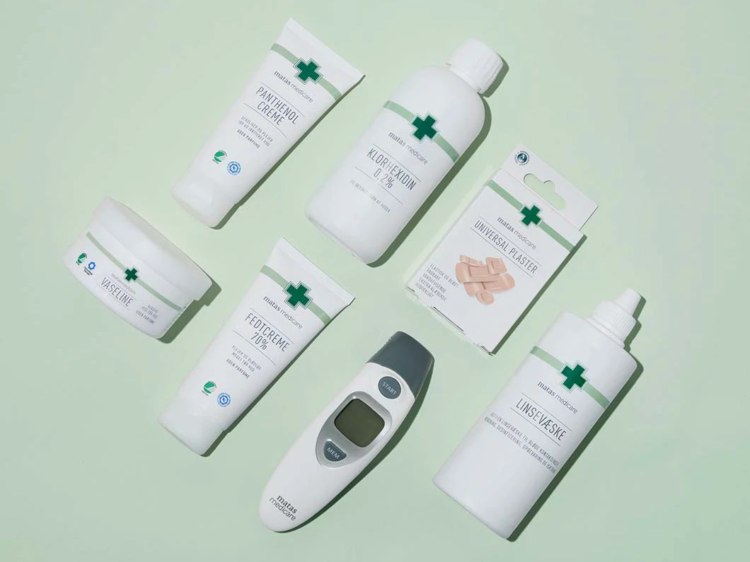 Matas Medicare-produkter ligger fladt på lysegrøn baggrund: panthenolcreme, klorhexidin, universalplastre, vaseline, fedtcreme, linsevask og et digitalt termometer.