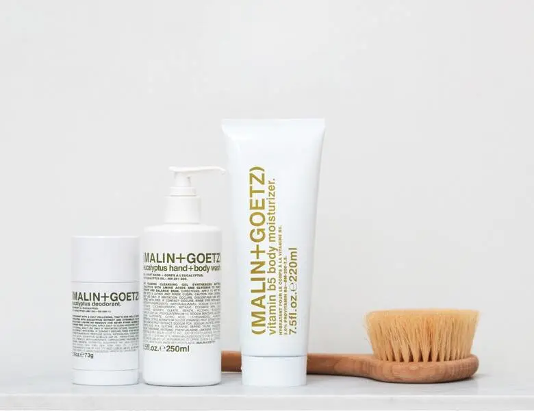 Her ses en hvid baggrund med en tørbørste og tre hvide Malin+Goetz produkter med brunlig skrift.