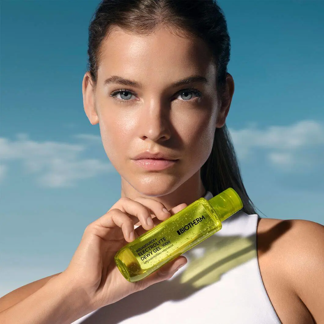 En kvindelig model med glødende hud og håret sat tilbage holder en grøn Biotherm Aquasource+ Electrolyte Dewy Gel-flaske op ved hagen. Hun ser direkte mod kameraet foran en blå himmel med lette skyer. Modellen bærer en hvid top, og billedet har et friskt og rent udtryk med fokus på fugtgivende hudpleje.