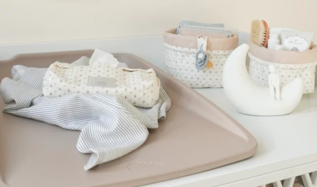 Pusleunderlag med stofble og vådservietter ligger på et puslebord, omgivet af små opbevaringskurve med babyplejeprodukter.