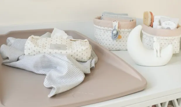 Pusleunderlag med stofble og vådservietter ligger på et puslebord, omgivet af små opbevaringskurve med babyplejeprodukter.