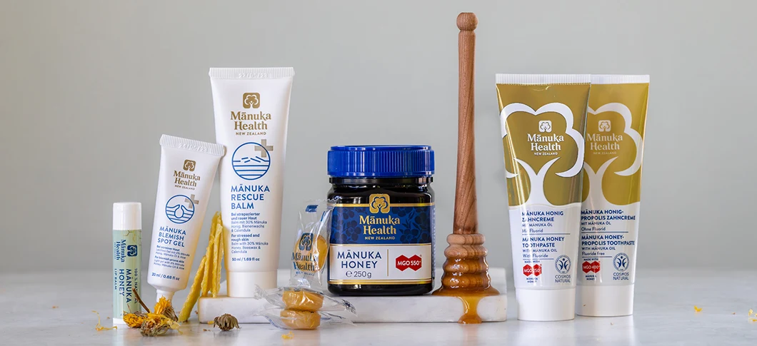 Manuka Health produkter står samlet på en lys overflade foran en neutral grå baggrund, herunder læbepomade, spot gel, rescue balm, manukahonning, mundskyl og tandpasta, sammen med gule blomster, små honningpastiller og en honningdypper.