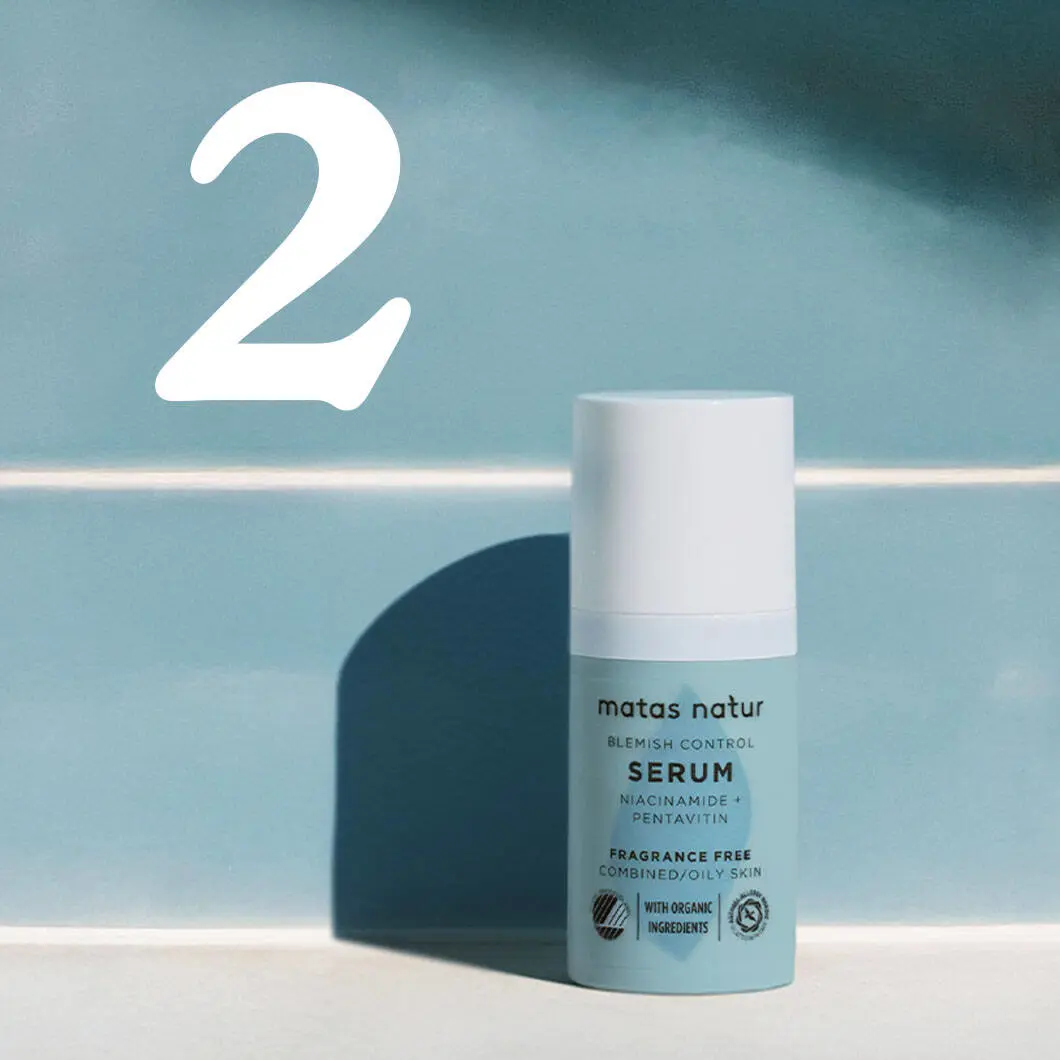 En lille flaske Matas Natur Blemish Control serum står på en lys overflade mod en blå baggrund. Flasken er lyseblå med hvidt låg. På etiketten står blandt andet ‘Serum’, ‘Niacinamide + Pentavitin’ og ‘Fragrance Free’. Til venstre ses et stort hvidt tal 2, som markerer andet trin i en hudplejerutine. Flasken kaster en blød skygge bag sig.