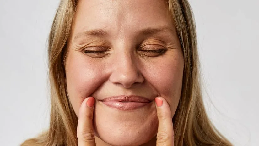 Nærbillede af en smilende kvinde med lukkede øjne, der peger let på sine kinder med begge pegefingre mod lys baggrund.