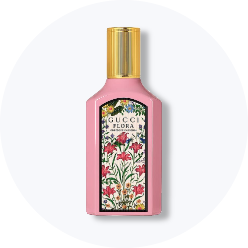 Her ses Gucci Flora Gorgeous Gardenia i en rosa parfumeflakon med guldfarvet låg. På forsiden ses en dekorativ etiket med farverige blomster og teksten “Gucci Flora Gorgeous Gardenia”. Flakonen står centralt mod en lys, rund baggrund.