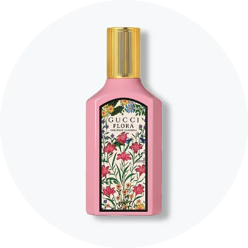 Her ses Gucci Flora Gorgeous Gardenia i en rosa parfumeflakon med guldfarvet låg. På forsiden ses en dekorativ etiket med farverige blomster og teksten “Gucci Flora Gorgeous Gardenia”. Flakonen står centralt mod en lys, rund baggrund.