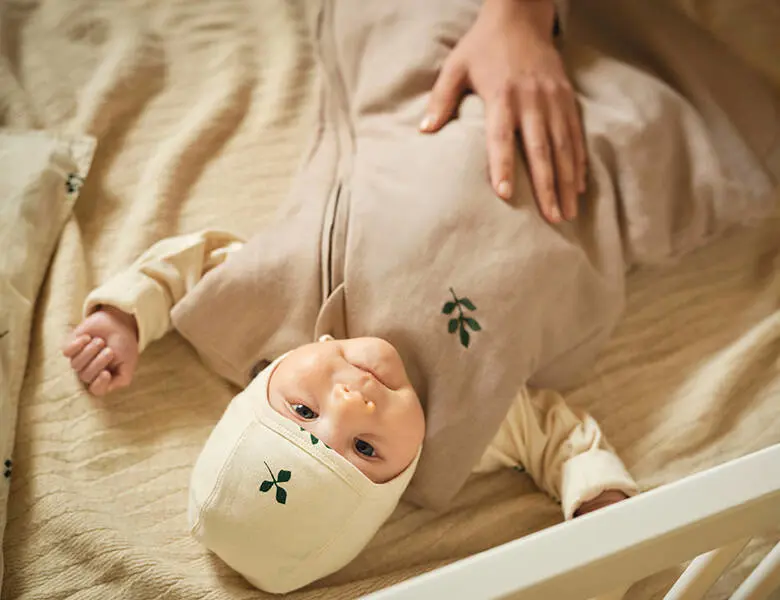 Baby med kyse og beige dragt med et enkelt lille grønt blad på kyse og tøj. Ligger på ryggen i tremmeseng og kigger op. En kvindes hånd hviler på barnets mave.