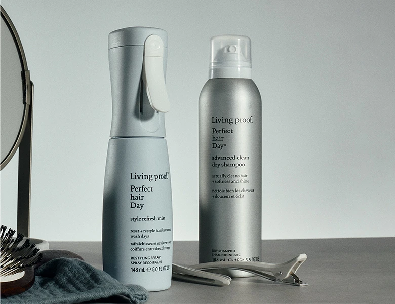 Her ses Living Proof Perfect Hair Day Style Refresh Mist og Perfect Hair Day Advanced Clean Dry Shampoo stående sammen med en hårbørste, hårklemmer og et spejl mod en lys baggrund.