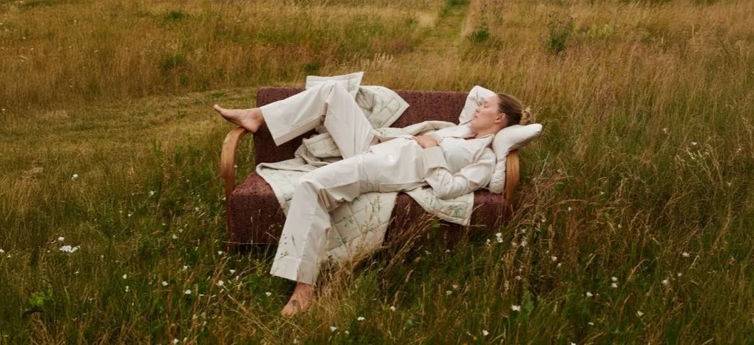 Her ses en kvinde, der ligger afslappet på en sofa med puder fra And Now You Sleep i en mark med højt græs iført lyst tøj.