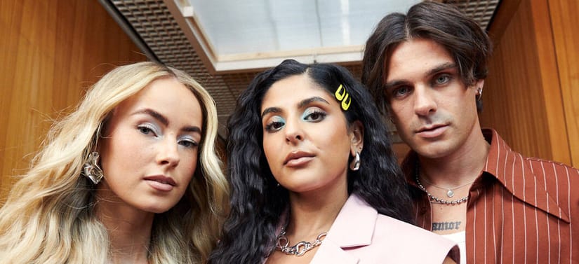 Her ses et billede med tre personer placeret tæt sammen og set forfra. De står foran en baggrund med træpaneler og loftdetaljer. Personerne har forskellige frisurer og makeup, og den midterste har et gult hårspænde i håret.