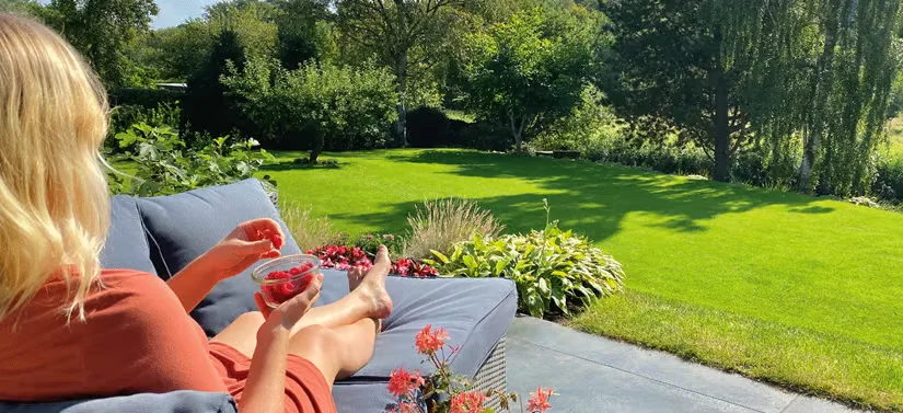Her ses en kvinde, som sidder i en havesofa med benene oppe. Hun spiser hindbær og kigger ud over sin flotte grønne have.