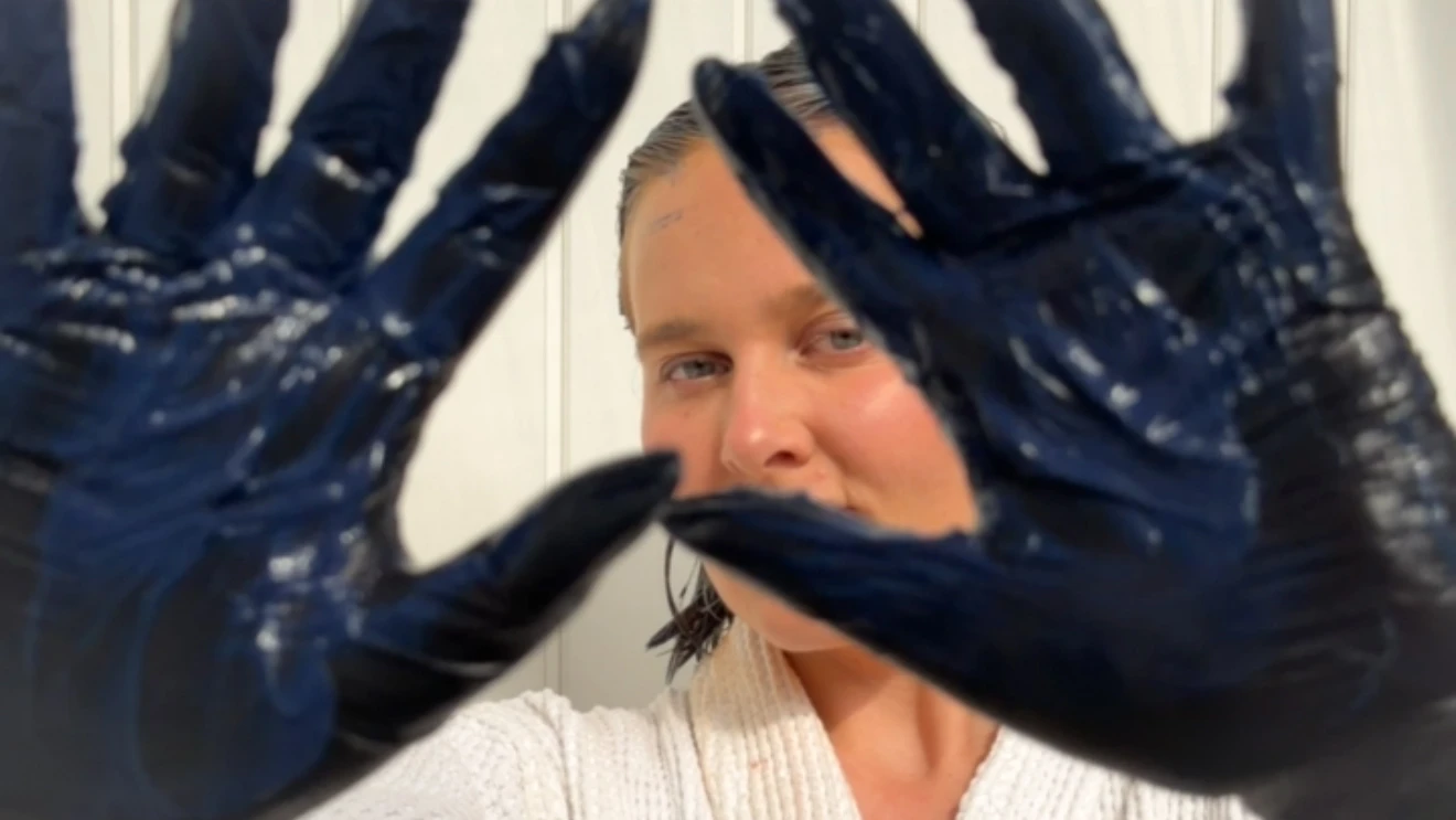 Person med vådt hår, smilende, holder hænder op i mørkeblå beskyttelseshandsker dækket af hårfarve.