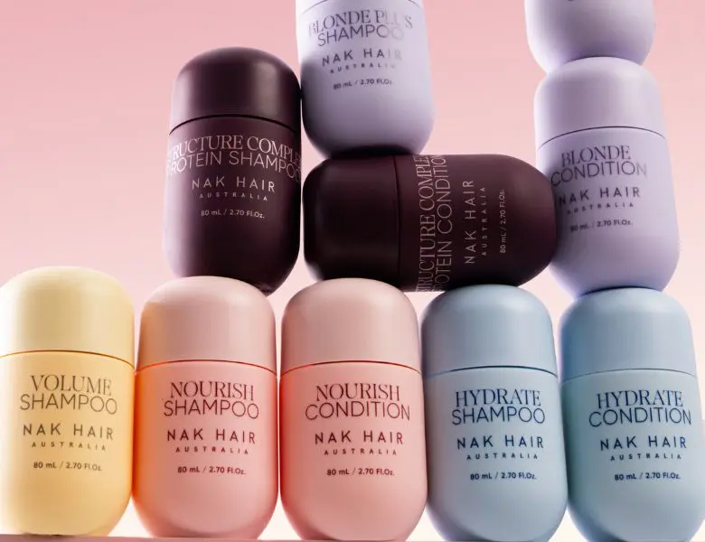 Her ses et billede med rejsestørrelser af hårplejeprodukter fra NAK Hair placeret mod en lyserød baggrund. Produkterne omfatter blandt andet “Volume Shampoo”, “Nourish Shampoo”, “Nourish Conditioner”, “Hydrate Shampoo”, “Hydrate Conditioner”, “Blonde Plus Shampoo”, “Blonde Conditioner”, “Structure Complex Protein Shampoo” og “Structure Complex Protein Conditioner”. Produkterne er arrangeret stablet i forskellige pastelfarver.