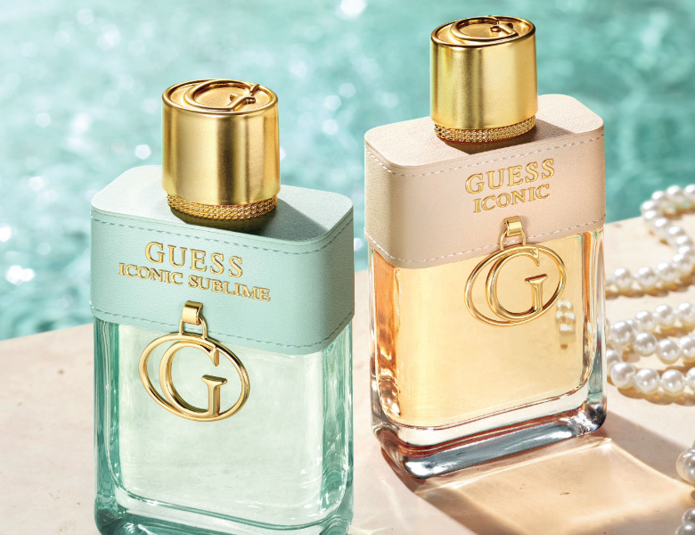 Her ses et billede med to parfumeflakoner fra GUESS placeret side om side mod en baggrund med vand. På flakonerne står teksten “GUESS ICONIC” og “GUESS ICONIC SUBLIME”. Flakonerne er i lyse grønne og gyldne nuancer med guldfarvede låg og et “G”-vedhæng.