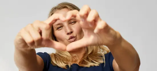 Her ses en pige, som laver et hjerte med hænderne. Baggrunden er grå.