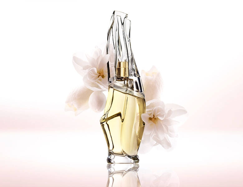 Her ses et billede af en parfumeflakon fra Donna Karan placeret centralt mod en lys baggrund. Flakonen har et asymmetrisk, transparent design med gyldent indhold og er omgivet af lyse blomster.