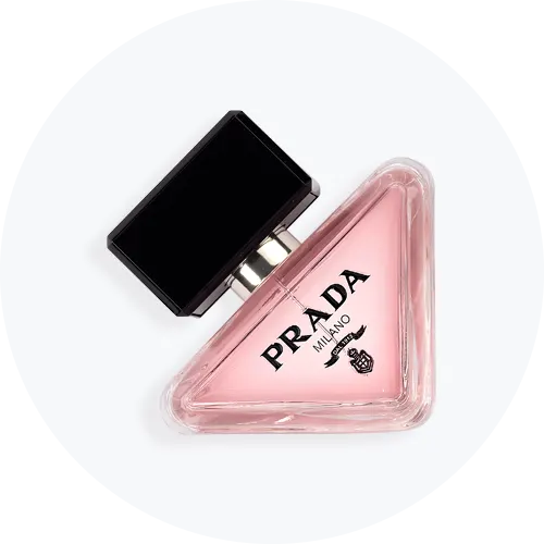 Her ses en trekantet Prada-parfumeflakon med rosa indhold og sort, geometrisk låg. På forsiden står “Prada Milano” sammen med brandets logo. Flakonen står centralt mod en lys, rund baggrund.