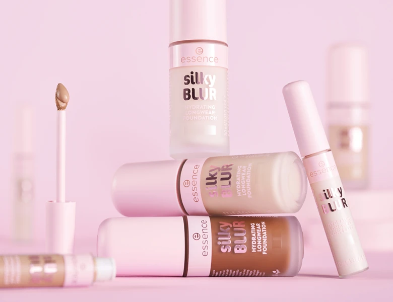 Her ses et billede med flere foundation-produkter fra essence placeret mod en lyserød baggrund. På produkterne står teksten “essence silky BLUR hydrating longwear foundation”. Der ses også en applikator med produkt på.