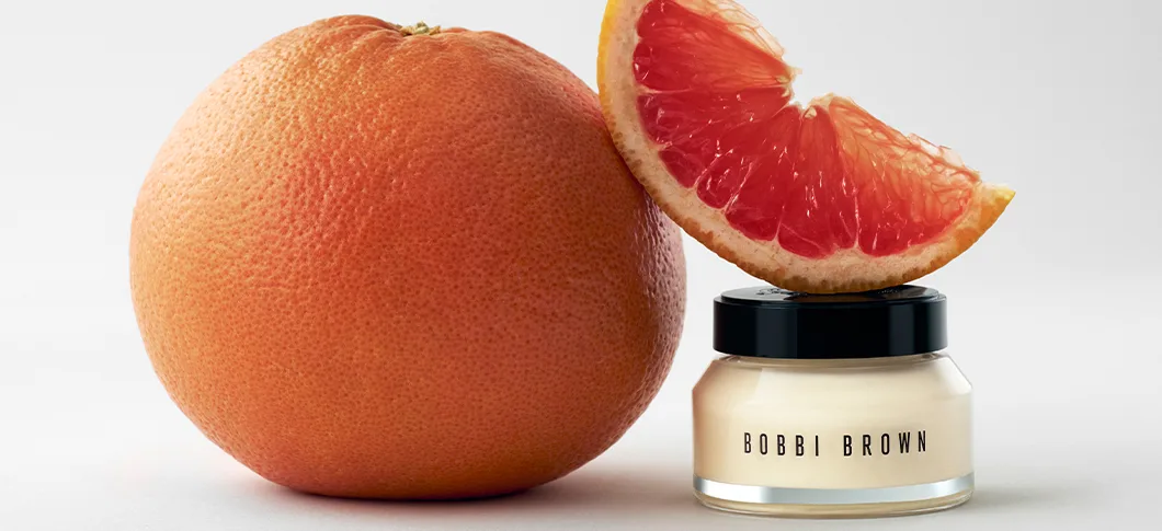 Her ses et billede med en krukke ansigtscreme fra Bobbi Brown placeret ved siden af en hel grapefrugt og en grapefrugtskive mod en lys baggrund. Kombinationen af hudplejeproduktet og citrusfrugten understreger et friskt og plejende udtryk.