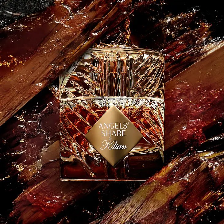 Klar glasparfumeflaske, Angels' Share by Kilian, med ravfarvet indhold, omgivet af flydende mørkebrune strøg.