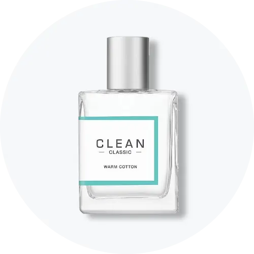 Her ses CLEAN Classic Warm Cotton i en transparent parfumeflakon med sølvfarvet låg. På forsiden ses en hvid etiket med turkis kant og teksten “CLEAN Classic – Warm Cotton”. Flakonen står centralt mod en lys, rund baggrund.