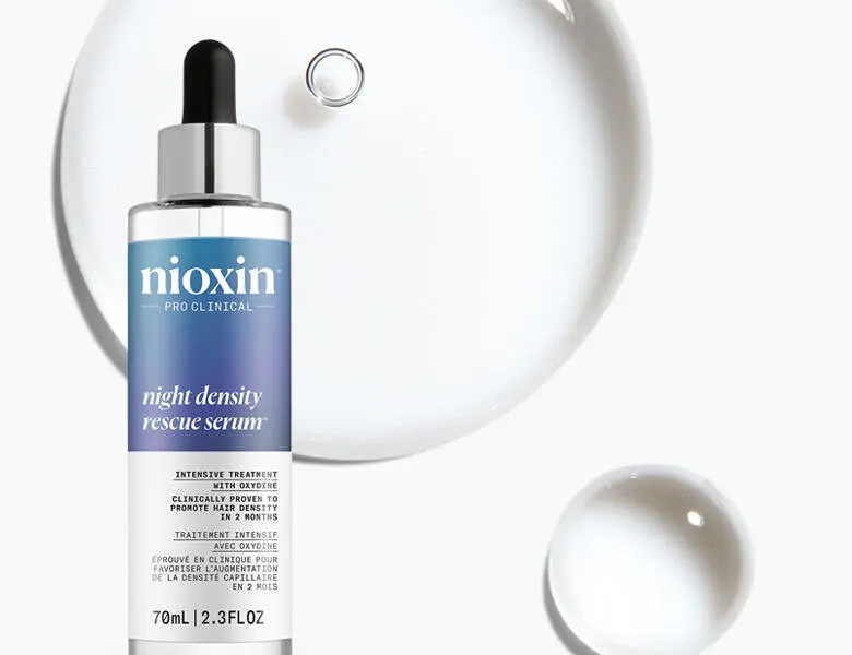 Her ses et billede af et hårserum fra Nioxin placeret mod en lys baggrund med gennemsigtige dråbeformer. På flasken står teksten “nioxin PRO CLINICAL night density rescue serum”. Produktet har pipette og blå detaljer på etiketten.