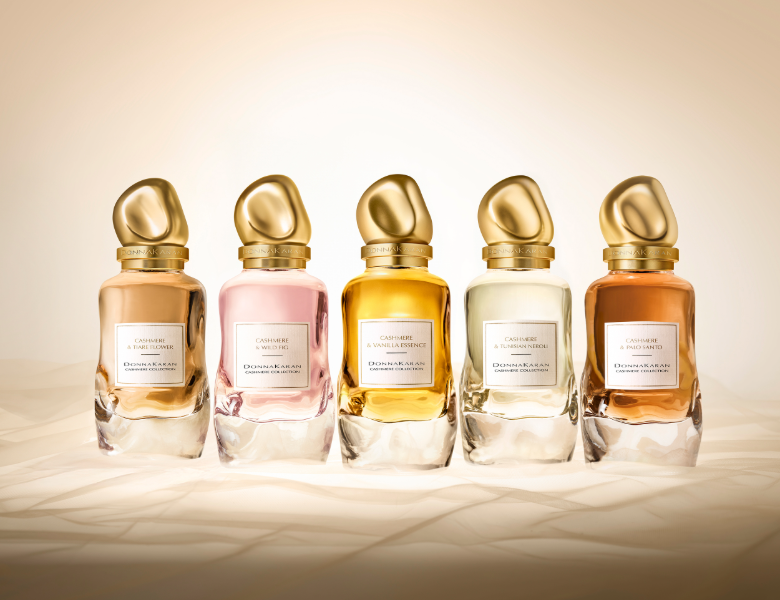 Her ses et billede med fem parfumeflakoner fra Donna Karan placeret side om side mod en lys, sandfarvet baggrund. På flakonerne står teksten “DONNA KARAN CASHMERE COLLECTION” samt forskellige varianter. Flakonerne er i gyldne, lyserøde og varme nuancer med organiske, guldfarvede låg.