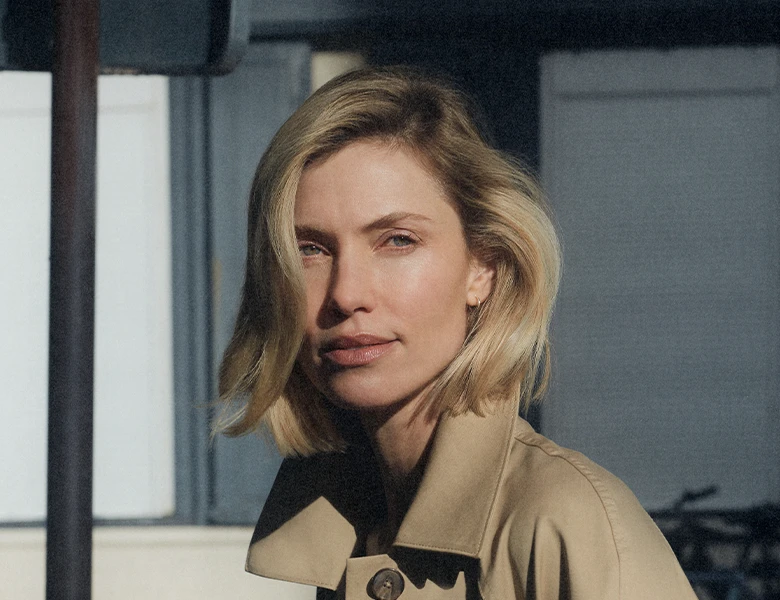 Her ses en person med kort, lyst hår iført en beige jakke. Personen står i sollys og kigger mod kameraet.