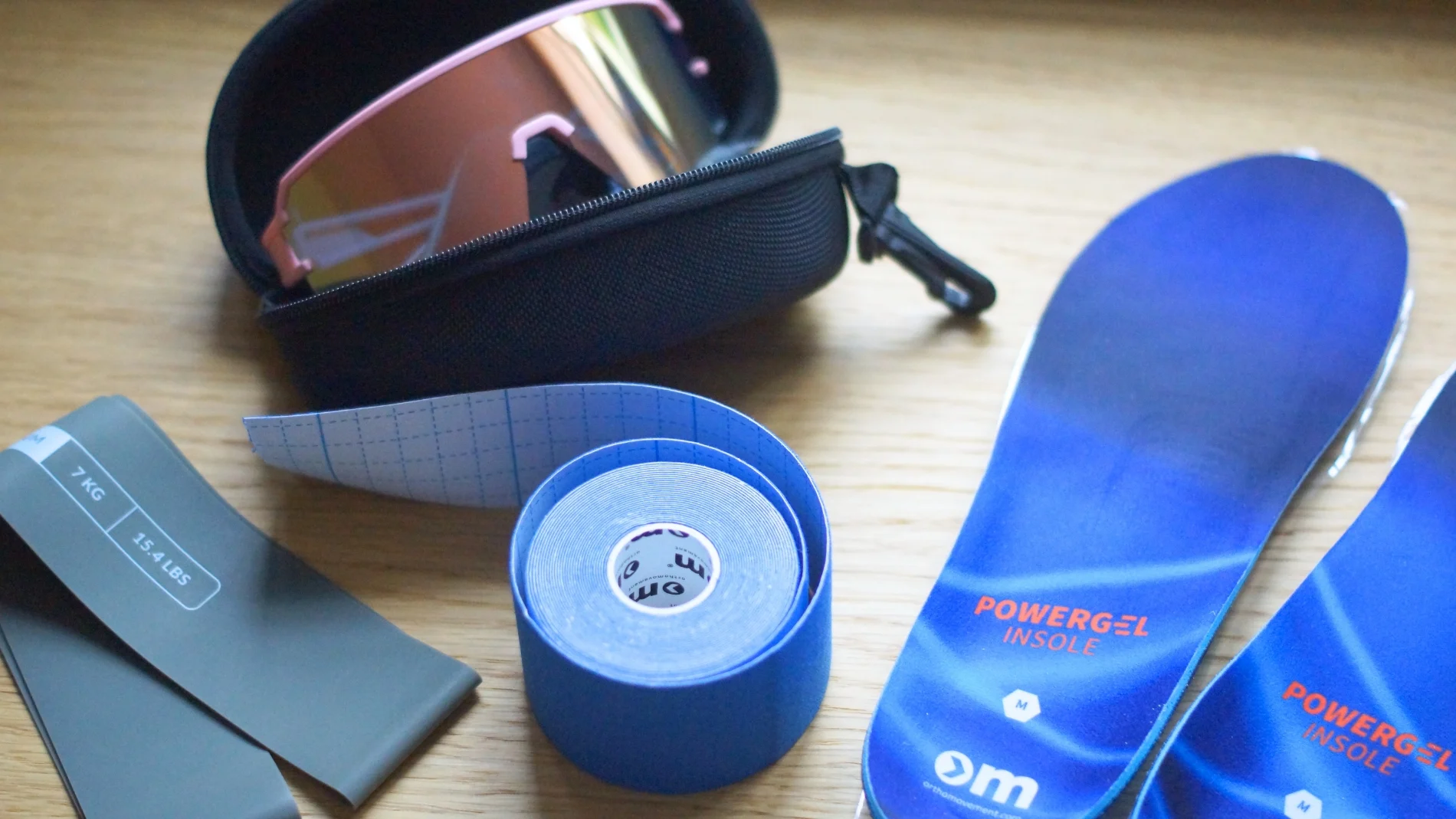 Forskelligt sportsudstyr: Lyserøde/orange solbriller i et sort etui, et gråt modstandsbånd mærket '7 KG 15.4 LBS', en rulle blå kinesiologitape med 'om orthamovement' logo, samt to blå 'POWERGEL INSOLE' indlægssåler i størrelse 'M' fra 'om orthamovement.com'.