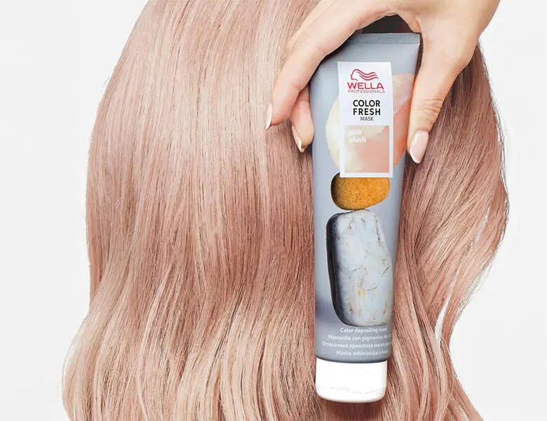 Her ses et billede af en person med lyst hår, der holder et hårprodukt fra Wella Professionals op mod håret. På tuben står teksten “COLOR FRESH MASK pink blush”. Produktet er i lyse nuancer med pink detaljer.