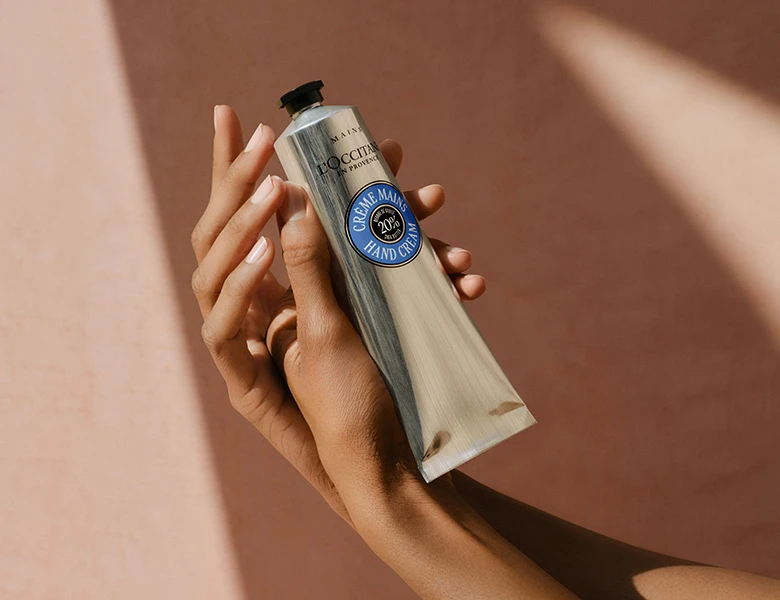Her ses en hånd, der holder en L’Occitane håndcreme foran en varm beige baggrund.