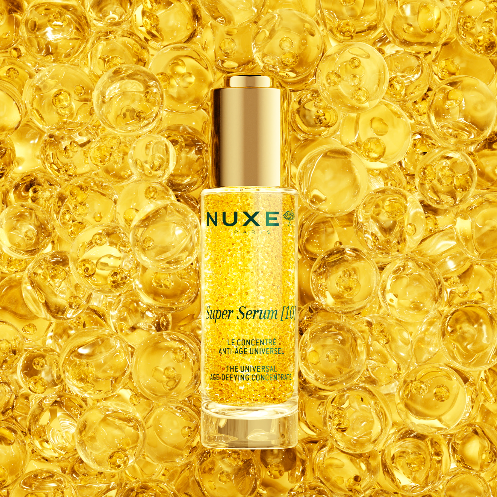 Nuxe Super Serum [10] med teksten 'LE CONCENTRÉ ANTI-ÂGE UNIVERSEL' og 'THE UNIVERSAL AGE-DEFYING CONCENTRATE', omgivet af gyldne oliekugler.