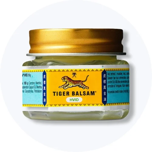 Tigerbalsam Hvid i lille glas med gul og blå etiket og gyldent låg på lys grå baggrund.