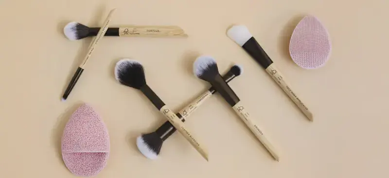 Forskellige makeupbørster med træskafter og sorte/hvide børster samt to lyserøde makeupsvampe.