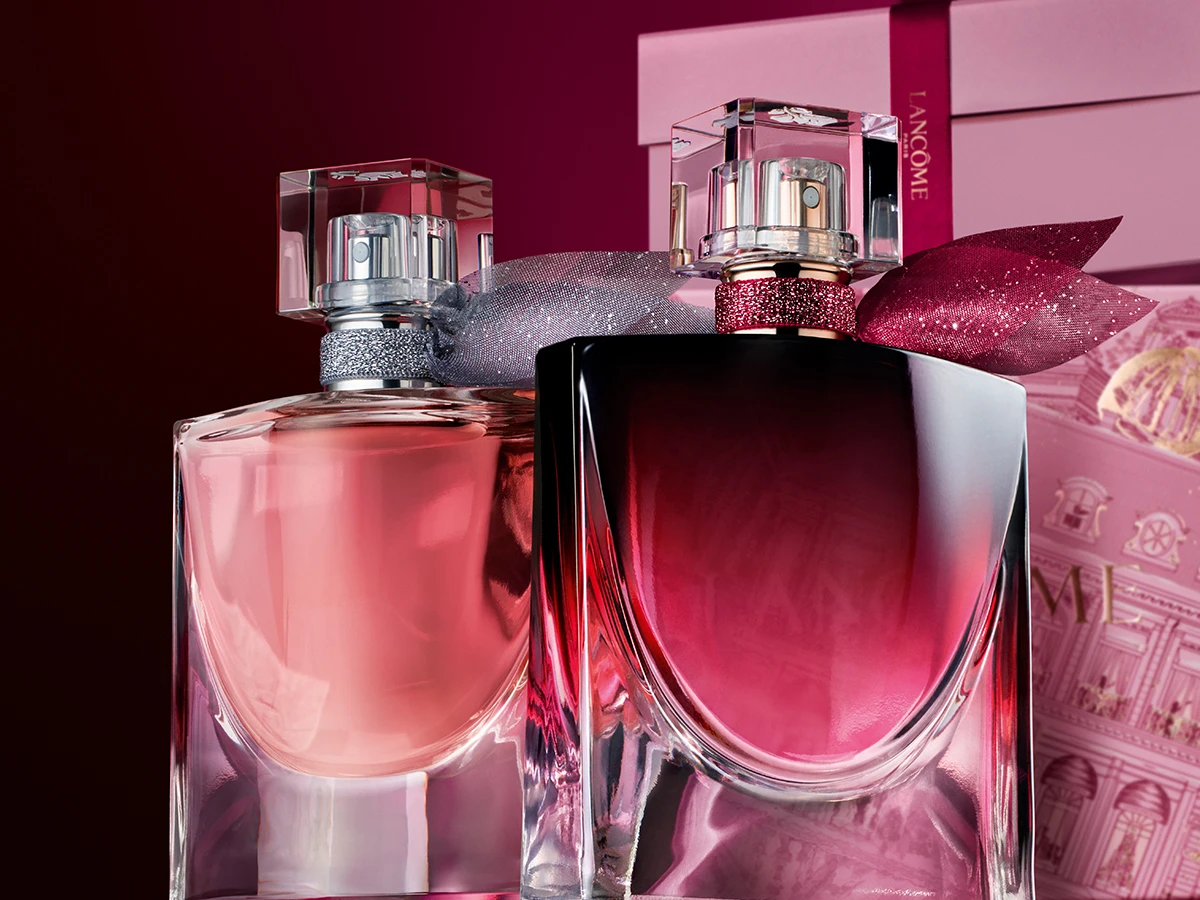 To Lancôme parfumeflasker, en lyserød og en mørk pink, med krystalhætter og glitrende bånd, foran lyserøde gaveæsker.