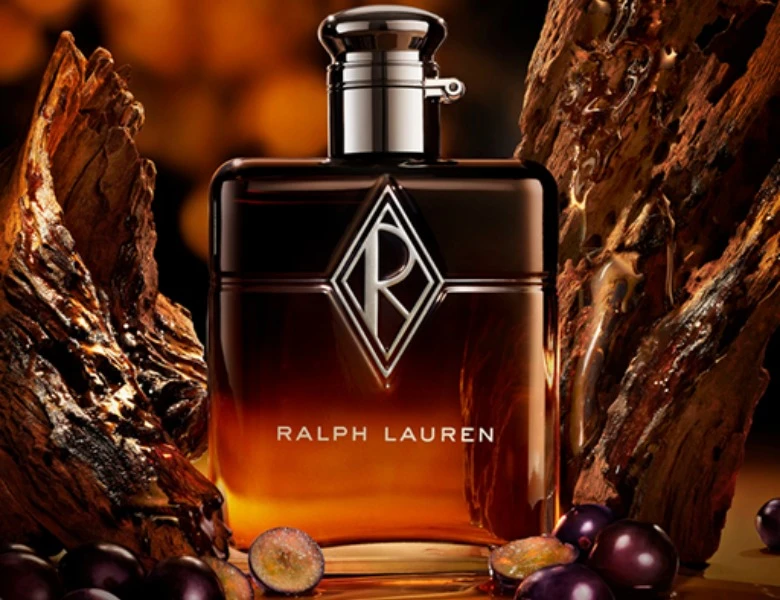 Her ses et billede af en parfumeflakon med teksten “RALPH LAUREN” på fronten. Flakonen er orange med et markeret logo på midten og placeret foran en mørk baggrund.