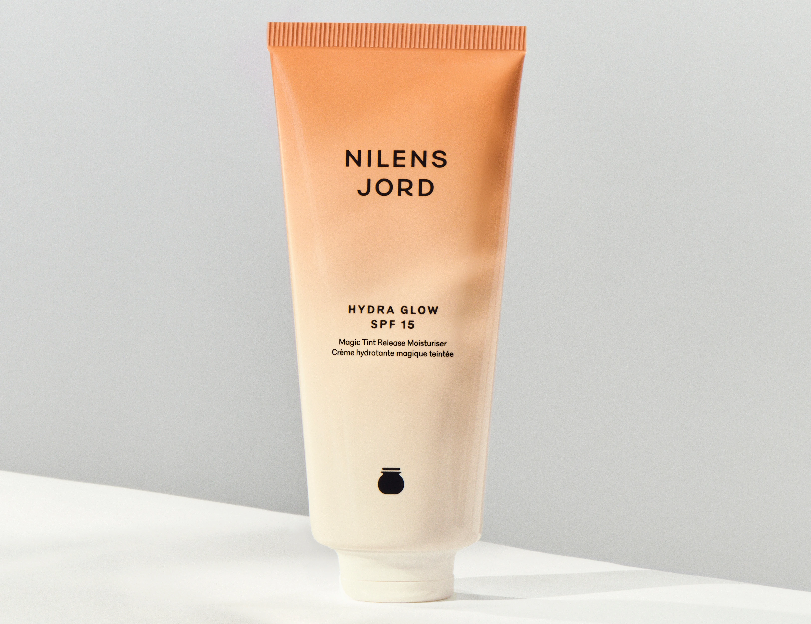 Her ses et billede af et hudplejeprodukt fra Nilens Jord placeret mod en lys baggrund. På tuben står teksten “NILENS JORD HYDRA GLOW SPF 15”. Produktet er i beige og cremefarvede nuancer.