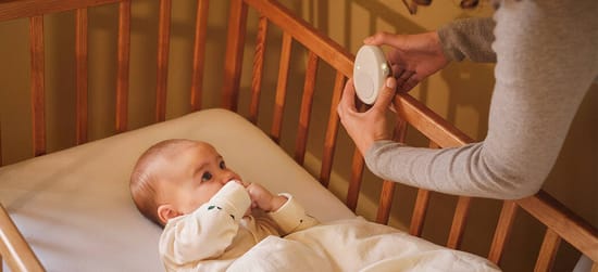 Her ses en baby, som ligger på en hvis madras i en babyseng af træ med trammer. En kvindes hånd kan ses nede i sengen sætte en white noise maskine på kanten.