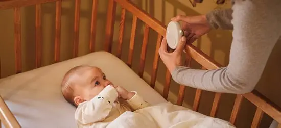 Her ses en baby, som ligger på en hvis madras i en babyseng af træ med trammer. En kvindes hånd kan ses nede i sengen sætte en white noise maskine på kanten.
