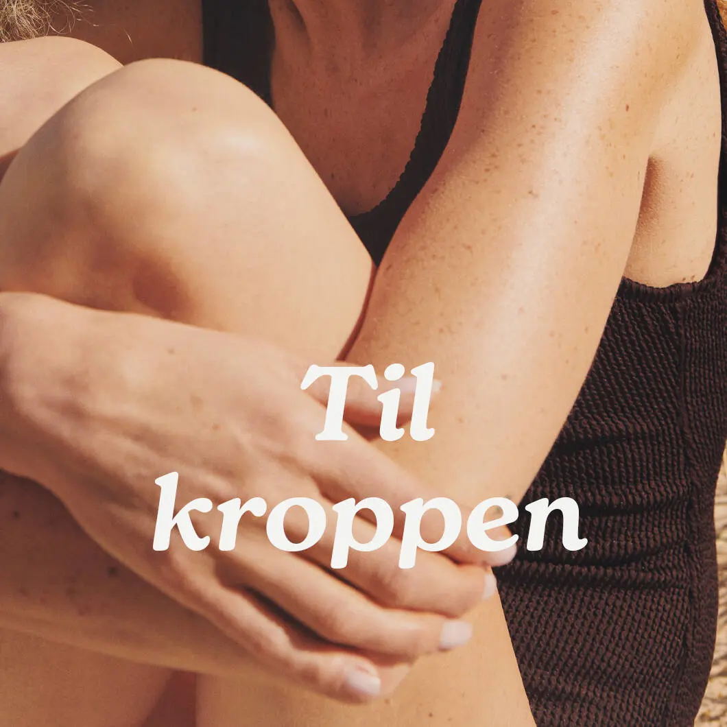 Nærbillede af en person i mørk badedragt, der sidder med armene omkring sine ben i varmt sollys. Midt på billedet står teksten: “Til kroppen”.