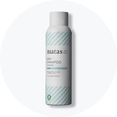 Her ses et billede af en dry shampoo fra Matas placeret centralt mod en lys, rund baggrund. På flasken står teksten “matas DRY SHAMPOO”. Flasken er i hvid med lyseblå stribede detaljer.