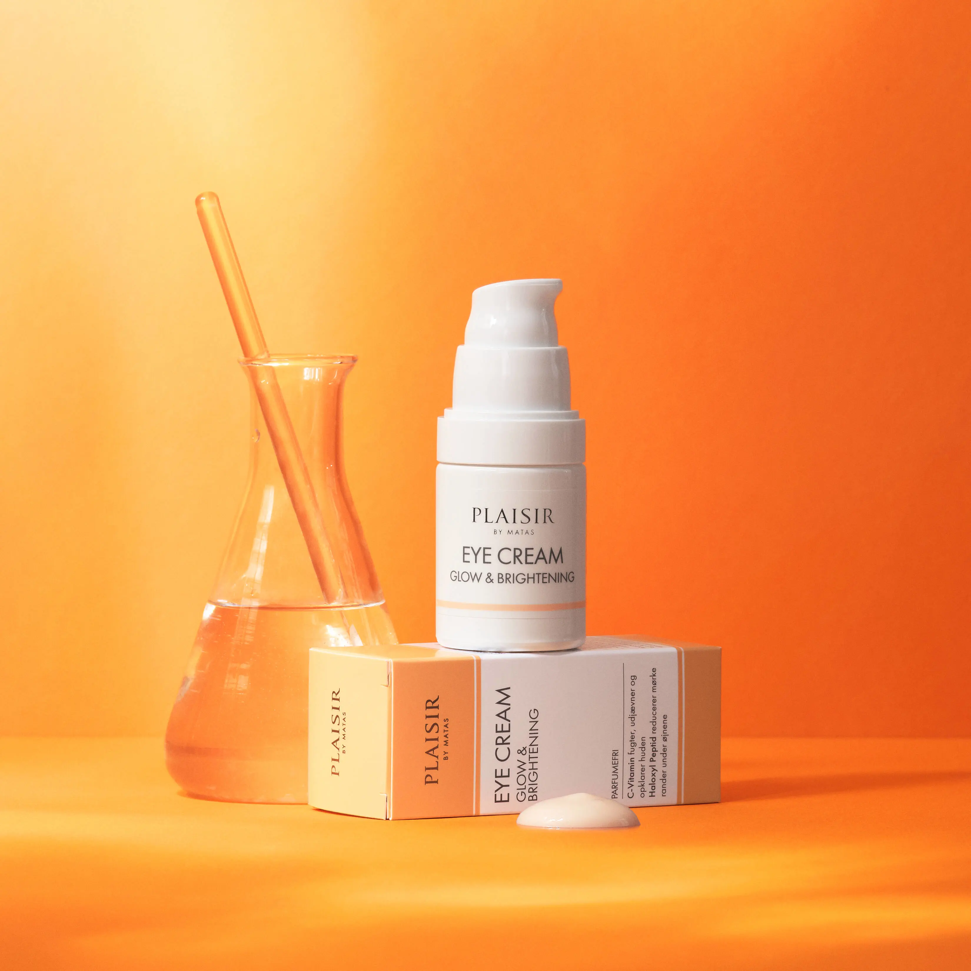 Produktbillede af øjencremen “Plaisir by Matas Eye Cream Glow & Brightening” på en varm orange baggrund. En hvid pumpeflaske står oven på den tilhørende produktæske i hvide og lyse orange farver. Ved siden af produkterne står en lille glasbeholder med klar væske og en glasstav. Foran æsken ligger en lille klat creme. Billedet har et rent og luksuriøst udtryk med fokus på fugtgivende og glødgivende hudpleje.