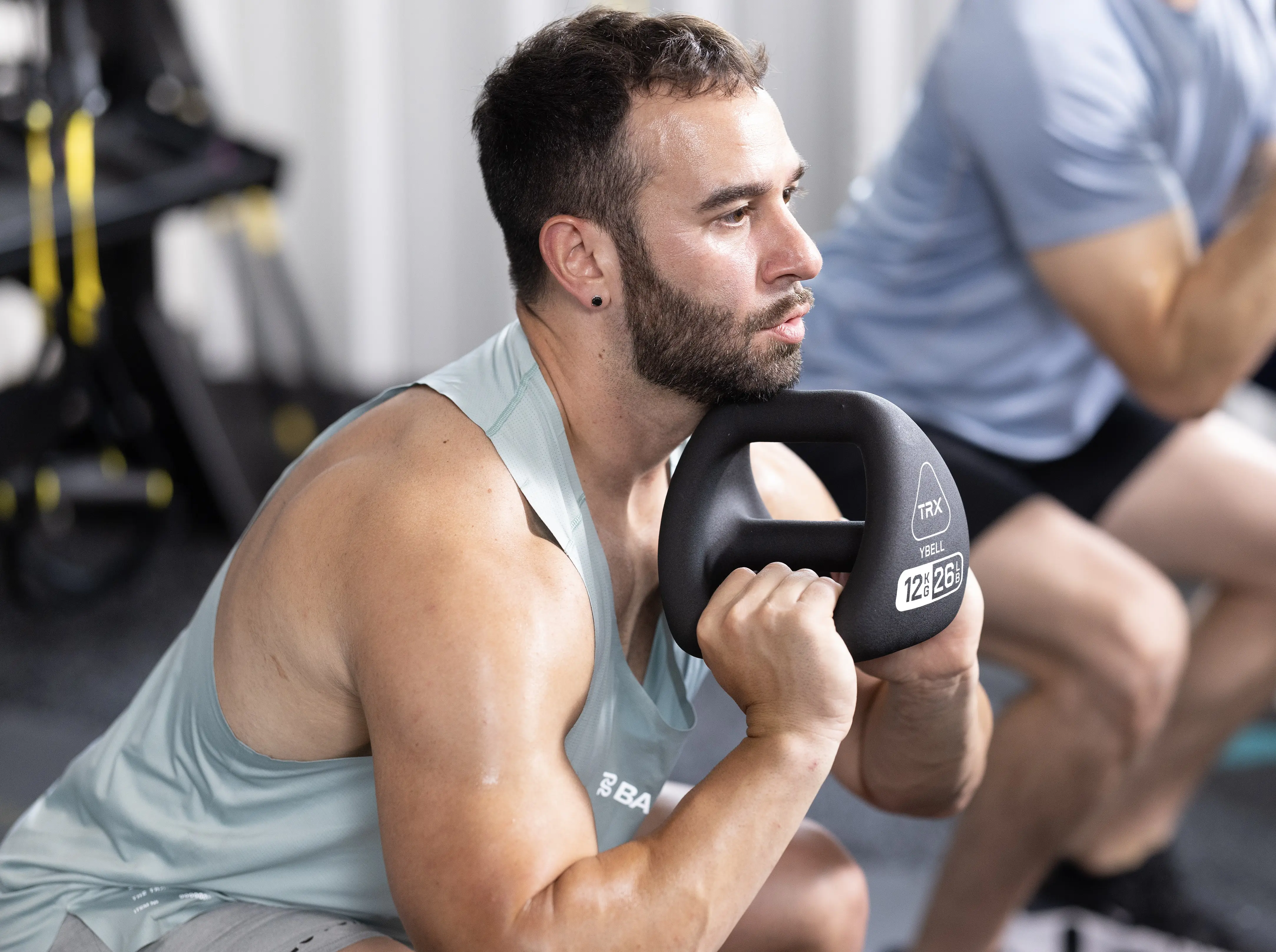 En mand i tanktop laver squat, mens han holder en sort kettlebell. Kettlebellen er mærket 'TRX YBELL 12kg 26lbs'.