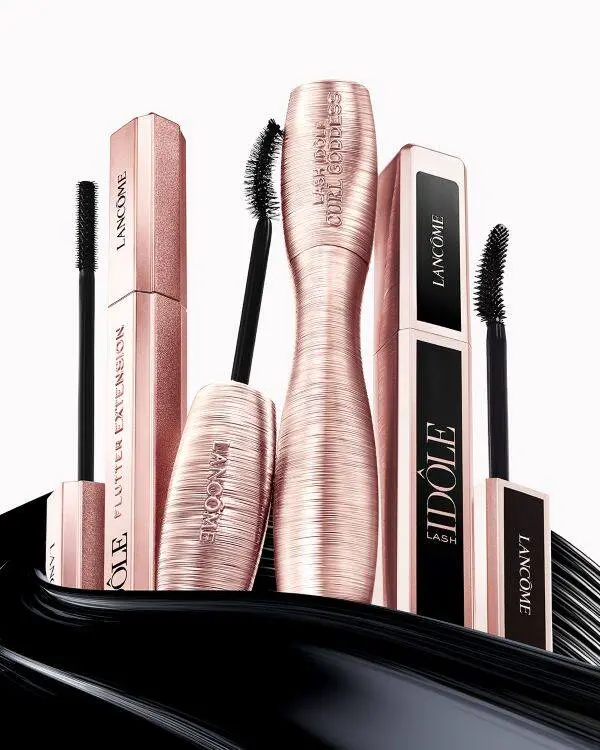 Et produktbillede viser flere Lancôme Lash Idôle-mascaraer i rosa metallic emballage arrangeret stående og åbne, så mascarabørsterne er synlige. Produkterne står foran en sort, bølgende mascaratekstur, som fremhæver den fyldige mascaraeffekt. Designet er elegant og moderne med fokus på lange, buede og definerede øjenvipper.